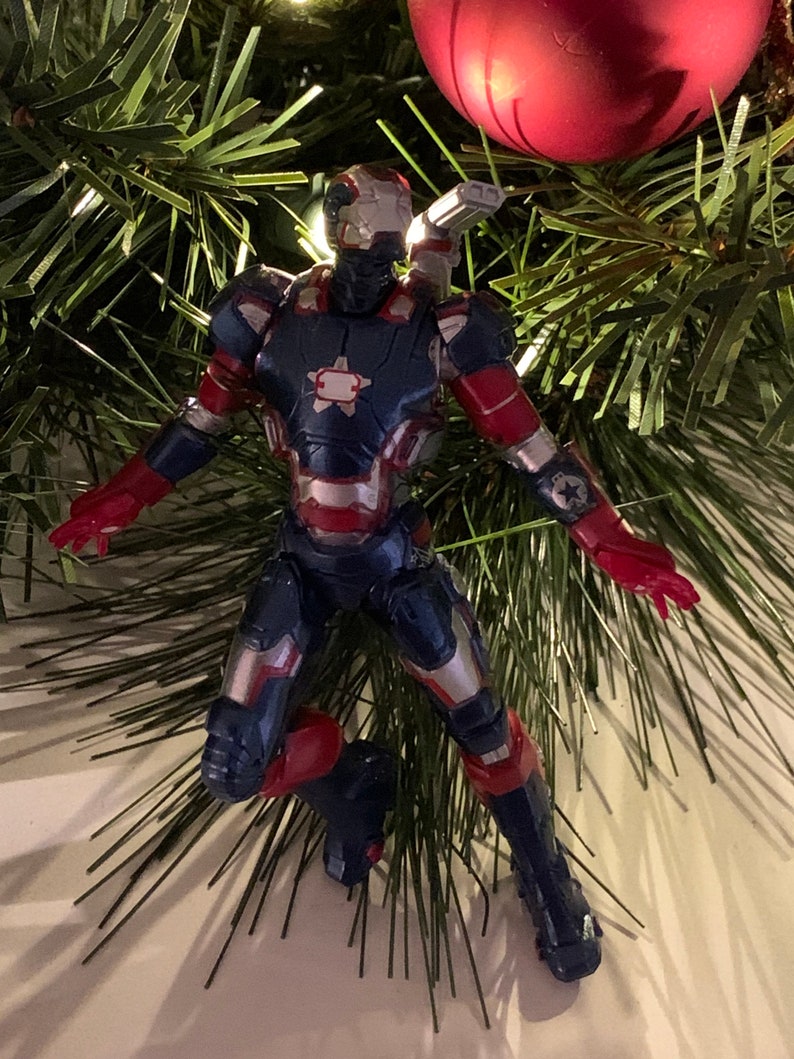 May include: A red, white and blue action figure of Iron Man in a flying pose. The figure is a Christmas ornament and is hanging from a Christmas tree.