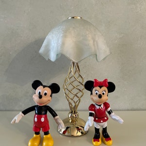 May include: A white lamp with a gold base and a white frosted glass shade.  Two plastic figurines of Mickey Mouse and Minnie Mouse are in front of the lamp. Mickey Mouse is wearing red shorts and yellow shoes. Minnie Mouse is wearing a red dress with white polka dots and yellow shoes.