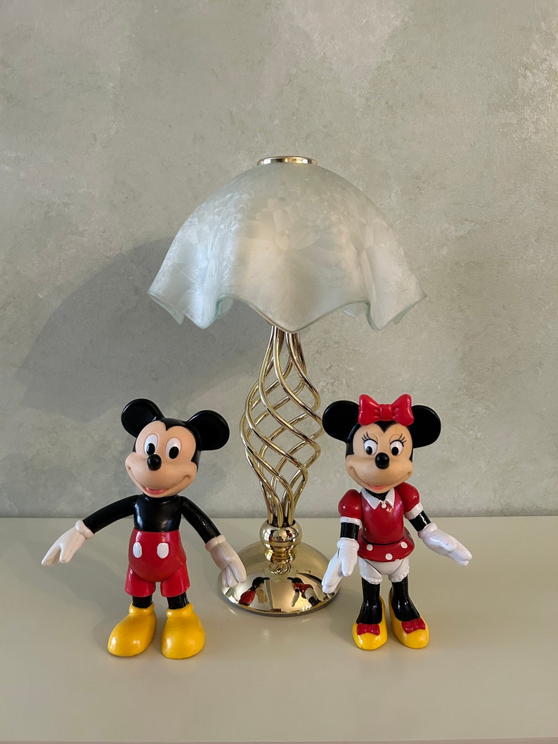 May include: A white lamp with a gold base and a white frosted glass shade.  Two plastic figurines of Mickey Mouse and Minnie Mouse are in front of the lamp. Mickey Mouse is wearing red shorts and yellow shoes. Minnie Mouse is wearing a red dress with white polka dots and yellow shoes.