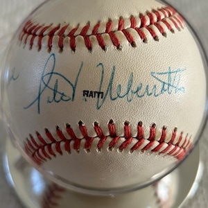 May include: A white baseball with red stitching and a blue signature that reads "Pete Rose" on the side. The baseball is in a clear display case.