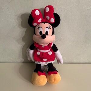 Op de afbeelding: Een rood en wit gestippeld pluche speelgoed van Minnie Mouse. Ze draagt een rode jurk met witte stippen en een rode strik met witte stippen. Ze heeft zwarte oren en een zwarte neus.
