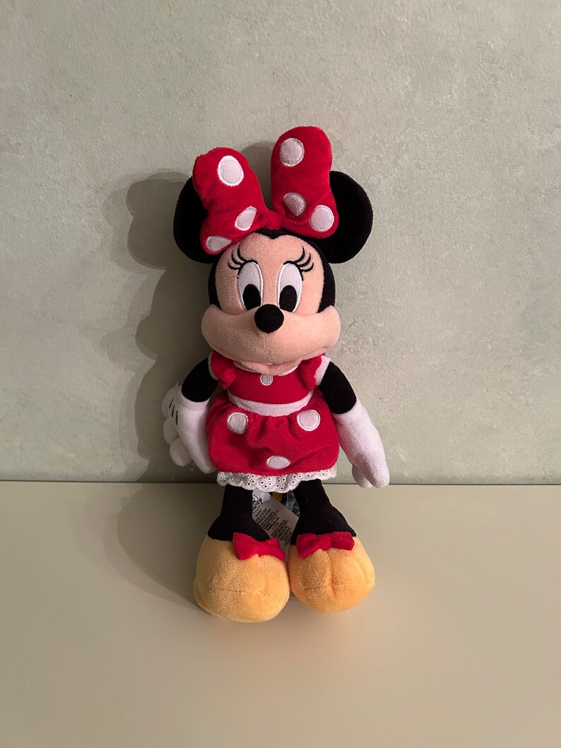 Op de afbeelding: Een rood en wit gestippeld pluche speelgoed van Minnie Mouse. Ze draagt een rode jurk met witte stippen en een rode strik met witte stippen. Ze heeft zwarte oren en een zwarte neus.