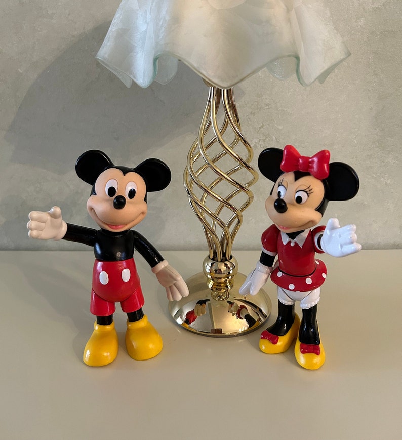 May include: Two plastic Disney character figurines, Mickey Mouse and Minnie Mouse. Mickey Mouse is wearing red shorts and yellow shoes. Minnie Mouse is wearing a red dress with white polka dots and yellow shoes. The figurines are standing on a white surface.