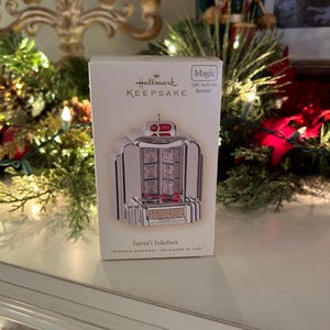 May include: A white Hallmark Keepsake box featuring a silver jukebox ornament labeled Santa's Jukebox. The box displays the text Magic Light, Sound, and Movement. The ornament has a coin slot, a selection panel with letters and numbers, and a central display area. The packaging is rectangular with branding at the top.