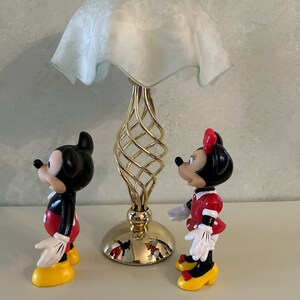 May include: A gold metal lamp with a white frosted glass shade.  Two plastic figurines of Mickey Mouse and Minnie Mouse stand in front of the lamp.