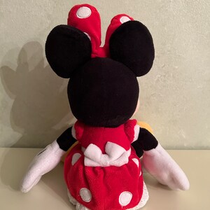 Op de afbeelding: Een rood en wit gestippeld pluche speelgoed van Minnie Mouse. Ze draagt een rode jurk met een witte strik en witte stippen. Ze heeft zwarte oren en een wit gezicht.