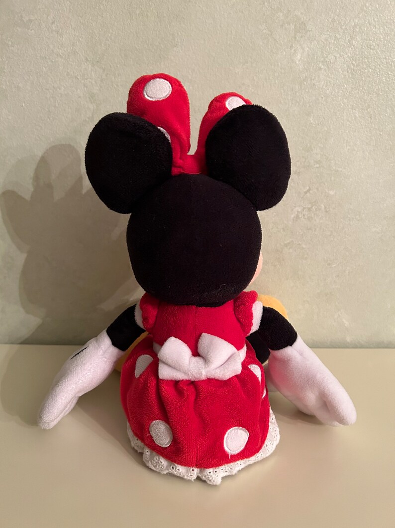 Op de afbeelding: Een rood en wit gestippeld pluche speelgoed van Minnie Mouse. Ze draagt een rode jurk met een witte strik en witte stippen. Ze heeft zwarte oren en een wit gezicht.