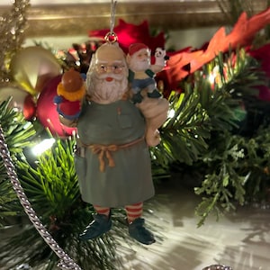 May include: A vintage Christmas ornament featuring a Santa Claus figure with a white beard and a green and red outfit. He is holding a small red and white figure and a small elf figure in his arms. The ornament is hanging from a silver wire.