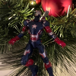 May include: A blue and red action figure of Iron Man in a flying pose, with a silver chest plate and a red star on the chest. The figure is on a Christmas tree with green pine needles and red and gold baubles.