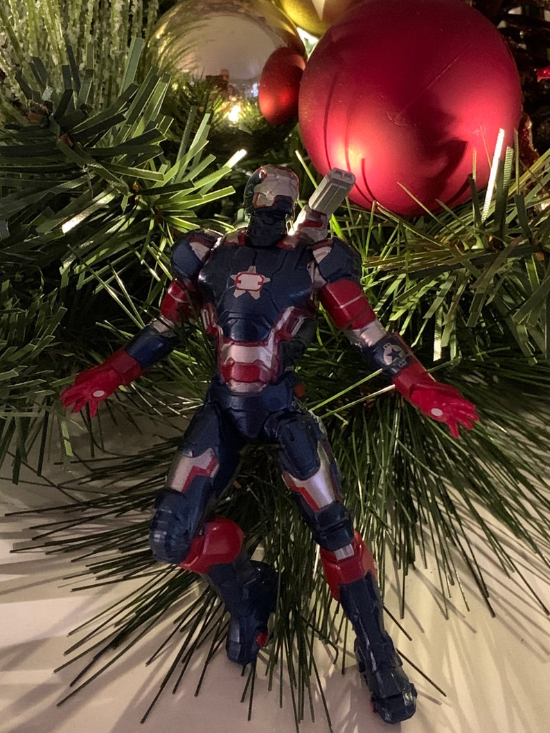 May include: A blue and red action figure of Iron Man in a flying pose, with a silver chest plate and a red star on the chest. The figure is on a Christmas tree with green pine needles and red and gold baubles.