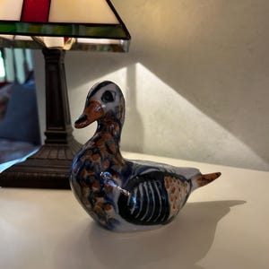May include: A ceramic duck figurine featuring a white base with hand-painted blue and orange spotted patterns. The duck has a smooth, glossy finish with a distinct orange beak and dark blue wing markings, captured in a side profile view.