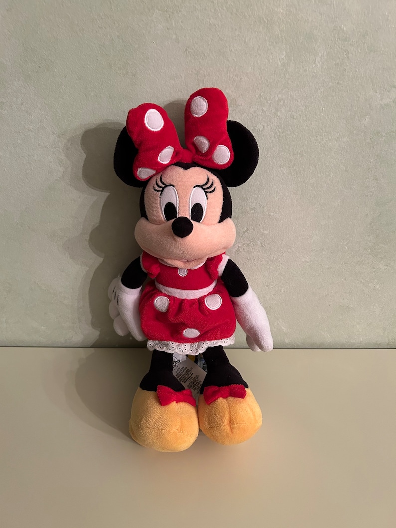 Op de afbeelding: Een rood en zwart pluche speelgoed van Minnie Mouse, die een rode jurk draagt met witte stippen en een rode strik met witte stippen.