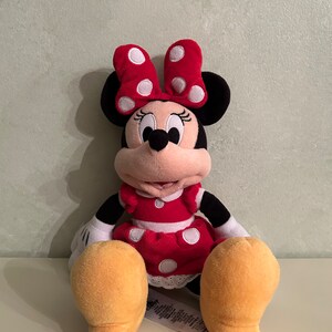Op de afbeelding: Een rood en wit gestippeld pluche speelgoed van Minnie Mouse. Ze draagt een rode jurk met witte stippen en een rode strik met witte stippen. Ze heeft zwarte ogen en een zwarte neus. Ze zit met haar benen gekruist en haar armen langs haar zijde.