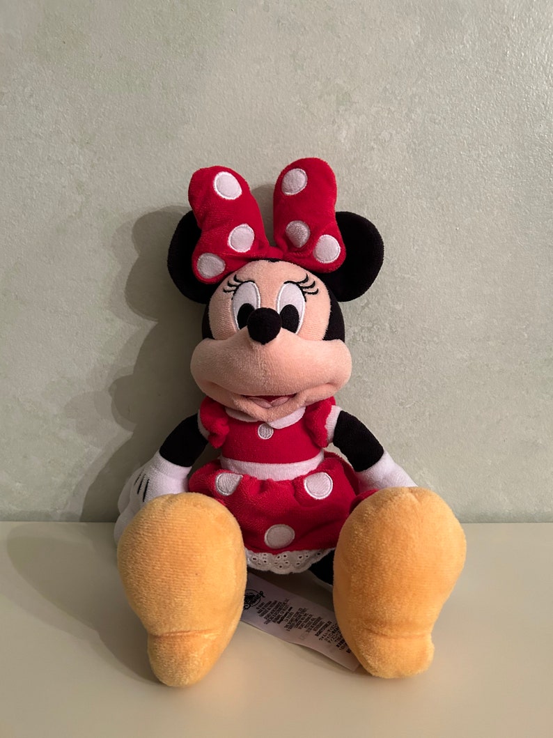 Op de afbeelding: Een rood en wit gestippeld pluche speelgoed van Minnie Mouse. Ze draagt een rode jurk met witte stippen en een rode strik met witte stippen. Ze heeft zwarte ogen en een zwarte neus. Ze zit met haar benen gekruist en haar armen langs haar zijde.