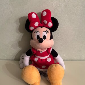 Op de afbeelding: Een pluche speelgoed van Minnie Mouse die een rode jurk draagt met witte stippen en een rode strik met witte stippen. Ze heeft zwarte oren en een zwarte neus. Haar ogen zijn zwart en haar mond is roze.