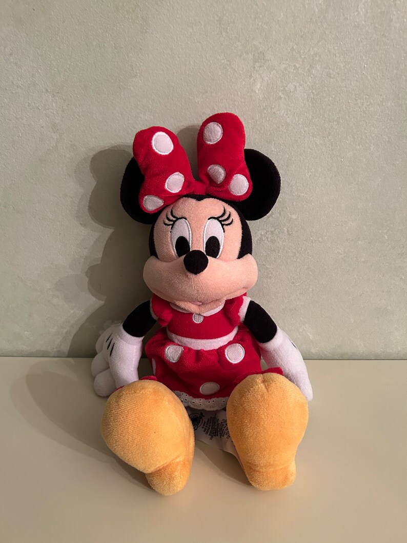 Op de afbeelding: Een pluche speelgoed van Minnie Mouse die een rode jurk draagt met witte stippen en een rode strik met witte stippen. Ze heeft zwarte oren en een zwarte neus. Haar ogen zijn zwart en haar mond is roze.