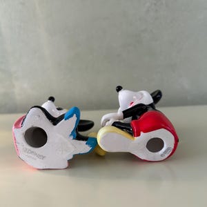 May include: Two ceramic Mickey Mouse figurines. One figurine has a red, black, and yellow outfit, and the other has a blue, pink, and white outfit. Both figurines are in a sitting position. The bottom of each figurine is stamped with "&copy;DISNEY JAPAN".