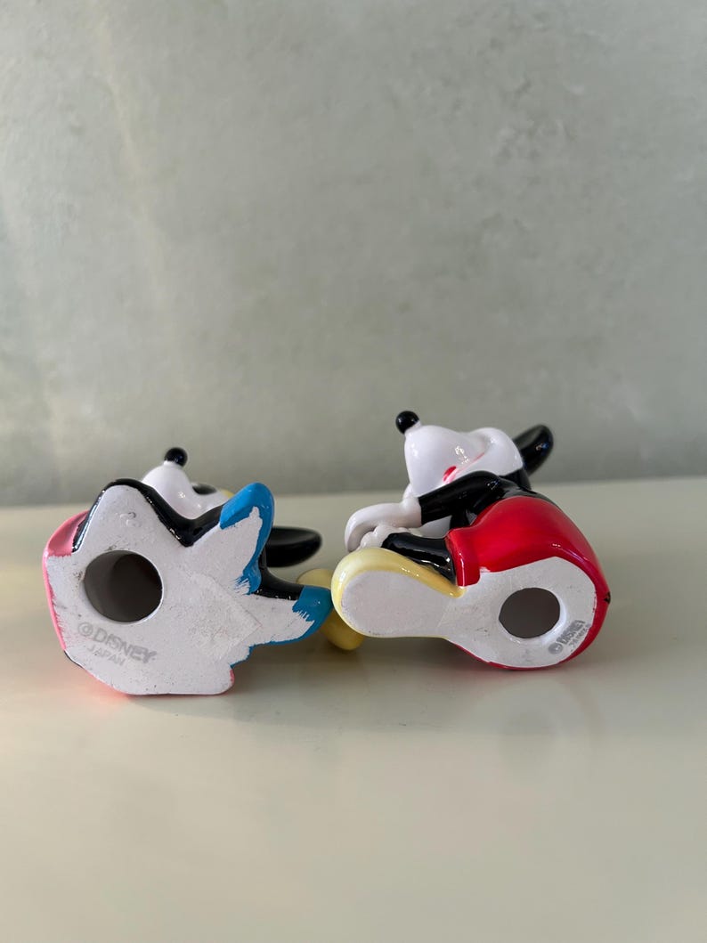 May include: Two ceramic Mickey Mouse figurines. One figurine has a red, black, and yellow outfit, and the other has a blue, pink, and white outfit. Both figurines are in a sitting position. The bottom of each figurine is stamped with "&copy;DISNEY JAPAN".