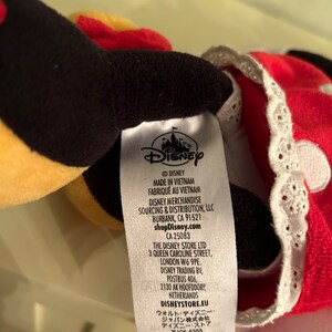 Op de afbeelding: Een wit label met zwarte tekst met details over de fabrikant, sourcing en distributie van een Disney pluche speelgoed. Het label bevat het Disney logo, de tekst "&copy;DISNEY", "MADE IN VIETNAM", "FABRIQU&Eacute; AU VIETNAM", "DISNEY MERCHANDISE SOURCING & DISTRIBUTION, LLC", "BURBANK, CA 91521", "shopDisney.com", "CA 25083", "THE DISNEY STORE LTD", "3 QUEEN CAROLINE STREET, LONDON W6 9PE.", "DISNEY TRADING BV, POSTBUS 406, 2130 AK HOOFDDORP, NETHERLANDS", "DISNEYSTORE.EU", "ウォルト・ディズニー・ジャパン株式会社", "ディズニー・ストア", "〒105-6355", "東京都港区虎ノ門 1-23-1 虎ノ門ヒルズ", "DISNEYSTORE.JP".