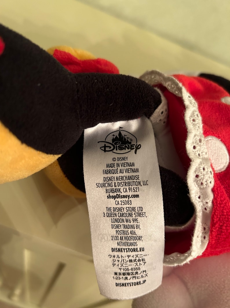 Op de afbeelding: Een wit label met zwarte tekst met details over de fabrikant, sourcing en distributie van een Disney pluche speelgoed. Het label bevat het Disney logo, de tekst "&copy;DISNEY", "MADE IN VIETNAM", "FABRIQU&Eacute; AU VIETNAM", "DISNEY MERCHANDISE SOURCING & DISTRIBUTION, LLC", "BURBANK, CA 91521", "shopDisney.com", "CA 25083", "THE DISNEY STORE LTD", "3 QUEEN CAROLINE STREET, LONDON W6 9PE.", "DISNEY TRADING BV, POSTBUS 406, 2130 AK HOOFDDORP, NETHERLANDS", "DISNEYSTORE.EU", "ウォルト・ディズニー・ジャパン株式会社", "ディズニー・ストア", "〒105-6355", "東京都港区虎ノ門 1-23-1 虎ノ門ヒルズ", "DISNEYSTORE.JP".