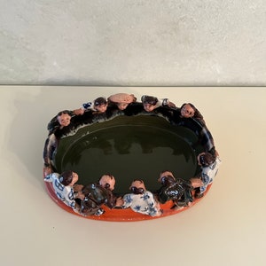 May include: A ceramic bowl with a green interior and an orange rim. The bowl is decorated with a circle of small figures in blue, white, and brown clothing. The figures are all facing inward, creating a circle around the bowl.