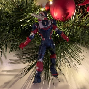 May include: A red, white and blue action figure of a superhero in a flying pose. The figure is posed as if it is hanging from a Christmas tree.
