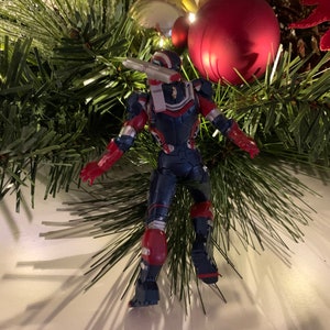 May include: A red, white and blue action figure of a superhero wearing a helmet and armour. The figure is standing on a white surface with a Christmas tree in the background.