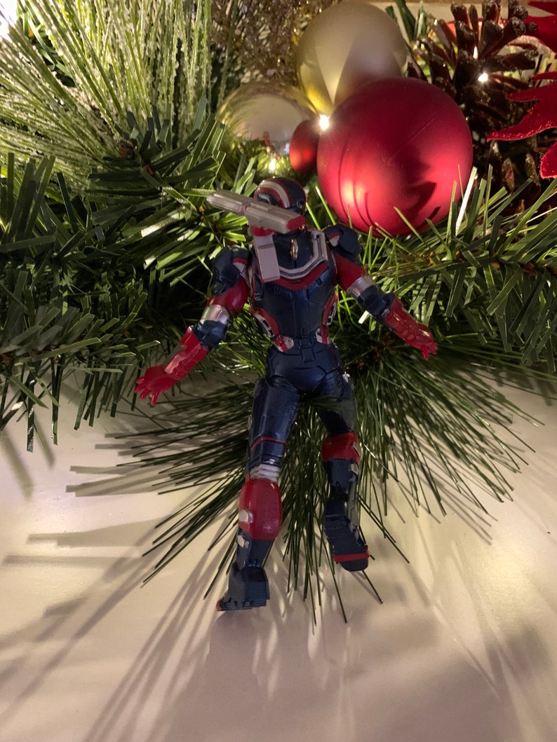 May include: A red, white and blue action figure of a superhero wearing a helmet and armour. The figure is standing on a white surface with a Christmas tree in the background.