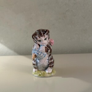 May include: A small ceramic figurine of a brown and white tabby cat standing on a green base. The cat has dark brown stripes, a light-colored chest, and is holding a blue and white patterned basket in its front paws with a pink bow on its back.