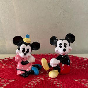 May include: Two ceramic figurines of Mickey and Minnie Mouse. Mickey is wearing red shorts, black shoes, and white gloves. Minnie is wearing a pink dress with white polka dots, blue shoes, and a yellow hat. Both are sitting on a red lace doily.