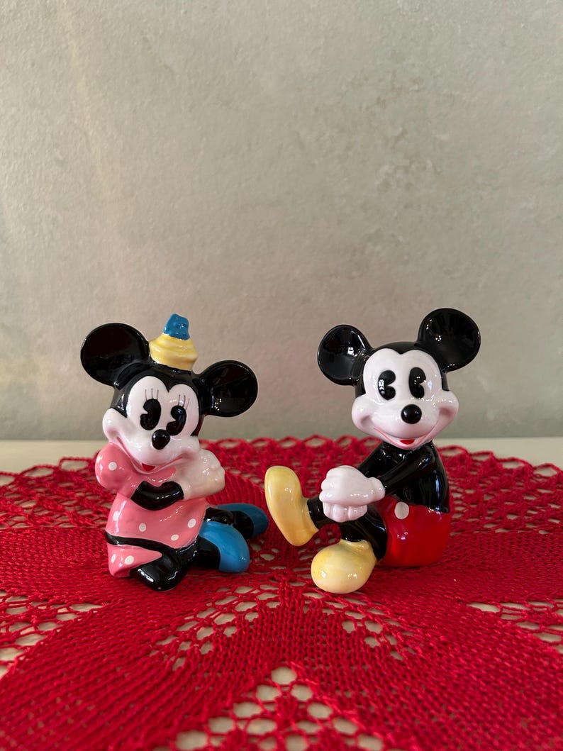 May include: Two ceramic figurines of Mickey and Minnie Mouse. Mickey is wearing red shorts, black shoes, and white gloves. Minnie is wearing a pink dress with white polka dots, blue shoes, and a yellow hat. Both are sitting on a red lace doily.
