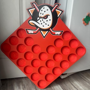 May include: A diamond-shaped red wooden display board featuring the Anaheim Ducks hockey team logo at the top. The board contains thirty-two circular cutouts arranged in a grid pattern, designed to hold small round items like hockey pucks or coins.