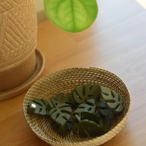 May include: A woven basket filled with several dark green monstera leaf-shaped objects. The basket sits on a wooden surface, with a potted plant and a large green leaf in the background. The objects are likely decorative.