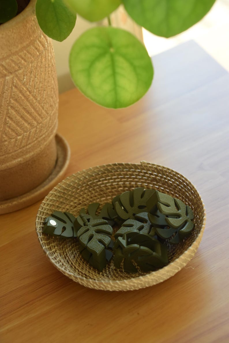 May include: A woven basket filled with several dark green monstera leaf-shaped objects. The basket sits on a wooden surface, with a potted plant and a large green leaf in the background. The objects are likely decorative.