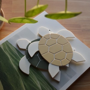 May include: Two decorative turtle figurines. One turtle has a tan shell and the other has a dark green shell. Both turtles have white flippers. The turtles are on a magazine with a green landscape image.