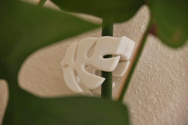 May include: A white, leaf-shaped plant support clip, designed to hold a plant stem. The clip is made of a solid, opaque material and is attached to a green stem. The background is a neutral, textured wall and green leaves.
