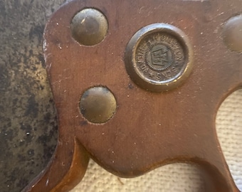Op de afbeelding: Close-up van een vintage handzaag. De zaag heeft een donkerbruine houten handgreep met messing klinknagels en een cirkelvormig embleem. Het zaagblad is van donker, getextureerd metaal. De handgreep heeft een gebogen ontwerp.