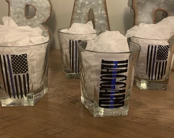May include: Four clear glass shot glasses with a blue line and American flag design. One glass has the word "THIN BLUE LINE" printed on it.