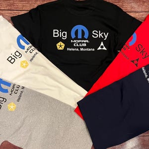 May include: A collection of t-shirts in various colors, including black, white, red, and gray. Each shirt features the text "Big Sky" and "MOPAR CLUB Helena, Montana" along with a logo. The shirts are displayed on a wooden surface.