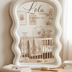 Peut inclure: Un miroir blanc aux bords ondulés avec des détails floraux et textuels gravés. Le miroir reflète un lit de bébé et comprend le nom "Lola", la date et l'heure. Une couverture pliée et une brosse sont posées sur une surface en bois.