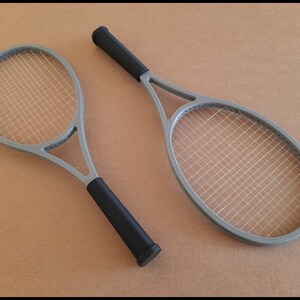 May include: Two gray tennis rackets with black grips are displayed against a neutral background. The rackets have a classic design with a gray frame and white stringing. The black grips provide a contrasting element to the rackets.