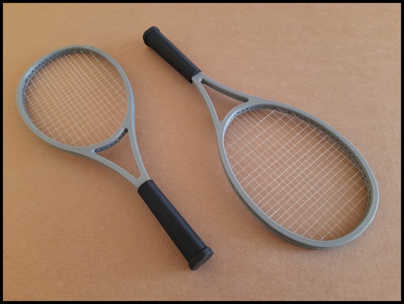 May include: Two gray tennis rackets with black grips are displayed against a neutral background. The rackets have a classic design with a gray frame and white stringing. The black grips provide a contrasting element to the rackets.