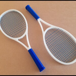May include: Two white tennis rackets with blue strings and blue handles are displayed against a tan background. The rackets are positioned at a slight angle, showcasing their oval-shaped heads and the intricate string patterns.