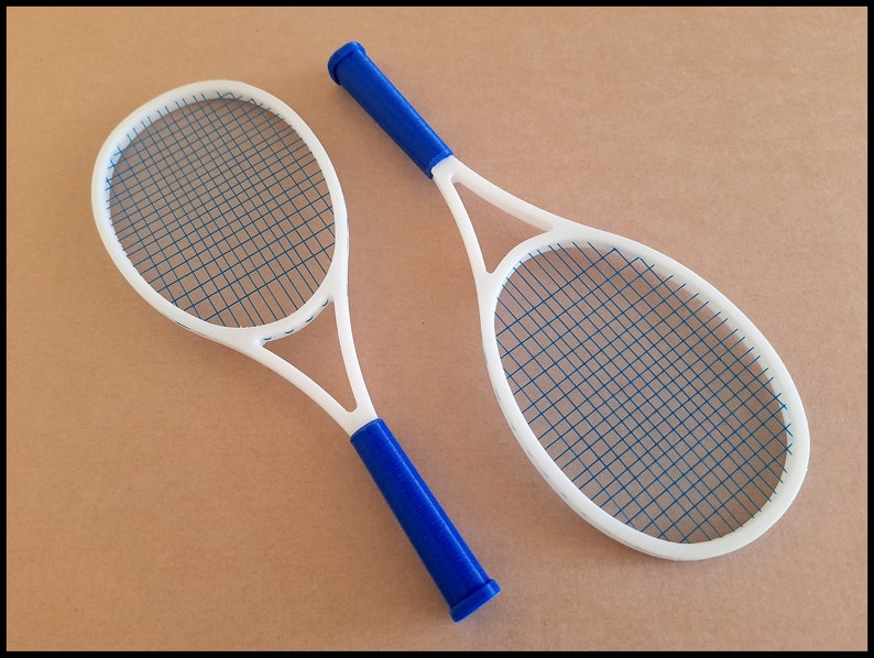 May include: Two white tennis rackets with blue strings and blue handles are displayed against a tan background. The rackets are positioned at a slight angle, showcasing their oval-shaped heads and the intricate string patterns.