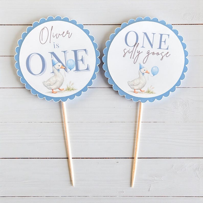 May include: Two round cake toppers with a scalloped blue border. One says "Oliver is ONE" and the other says "ONE silly goose". Each features a watercolor illustration of a goose wearing a party hat and holding a balloon. The toppers are on wooden sticks.