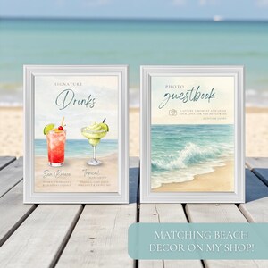 May include: Two framed signs with beach-themed watercolor art. One says "Signature Drinks" with illustrations of cocktails. The other says "Photo Guestbook" with a beach scene. The signs are on a wooden surface with a beach and ocean background.