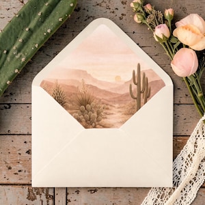 Pu&ograve; includere: Una busta color crema con un'illustrazione di un paesaggio desertico all'interno. L'illustrazione presenta cactus, montagne e un sole al tramonto nei toni del marrone e beige. La busta &egrave; su una superficie di legno con fiori e pizzo.