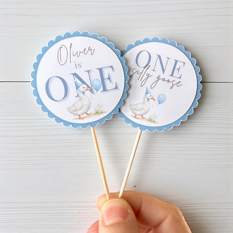 May include: Two round, scalloped-edge toppers with a light blue border. Each features a white goose wearing a party hat and holding a balloon. One says "Oliver is ONE", the other says "ONE silly goose".