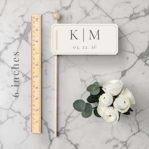 以下が含まれることがあります： イニシャル「K|M」と日付「03. 22. 26」が入ったクリーム色のウェディングケーキトッパー。トッパーの長さは約15cmで、白いバラと緑のブーケの隣にあります。木製の定規も画像にあります。