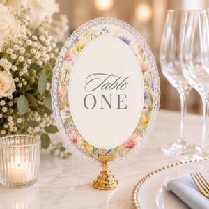 K&ouml;nnte beinhalten: Ein ovales Tischnummernschild mit einem Blumenmuster-Rand aus rosa, gelben und blauen Wildblumen, eingefasst in wei&szlig;e Spitze. Die Mitte ist cremefarben mit dem Text Table ONE in salbeigr&uuml;ner Serifenschrift auf einem goldfarbenen Metallst&auml;nder.